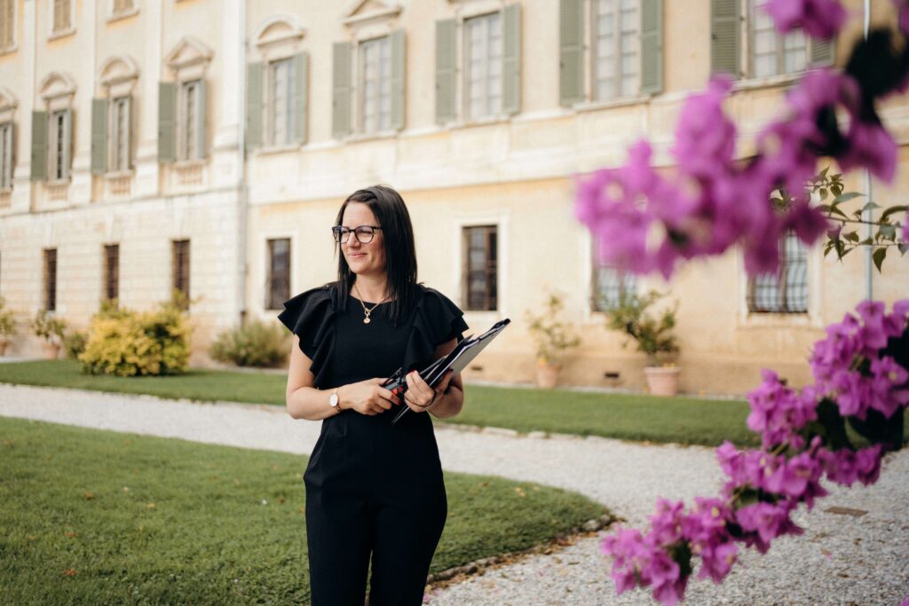 Eine Frau in schwarzem Outfit, die ein Haarglätteisen in der Hand hält, steht draußen in der Nähe eines historischen Gebäudes und lila Blumen - die perfekte Inspiration für jeden Hochzeitsplaner, der elegante Brautmomente in Apulien sucht.