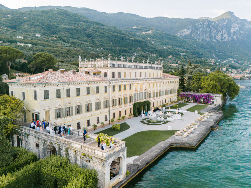 Luftaufnahme einer großen Villa am See in Apulien, Italien, mit Menschen, die sich zu einer Hochzeit inmitten atemberaubender Berge versammelt haben.