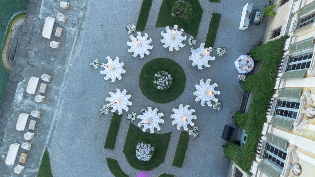 Luftaufnahme von runden Tischen, die für eine italienische Hochzeit im Freien auf einem gemusterten Hof aus Stein und Gras am atemberaubenden Comer See gedeckt sind.
