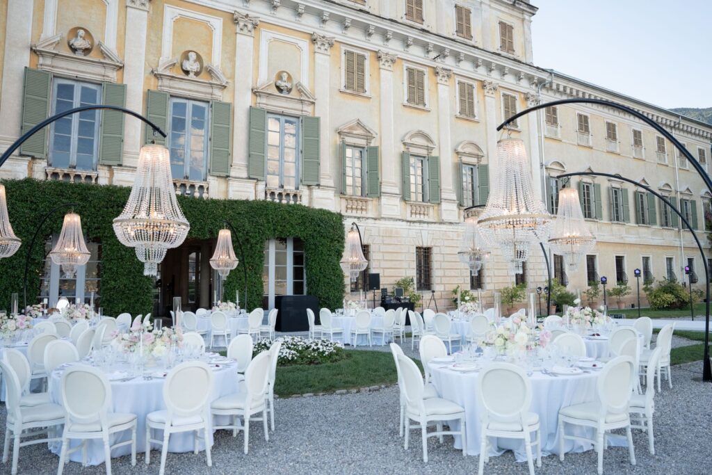 Elegante Hochzeitsfeier im Freien mit runden Tischen, weißen Stühlen und Kronleuchtern vor einem großen Gebäude - fachmännisch koordiniert von Hochzeitsplanerin Julia Weiß am wunderschönen Comer See.
