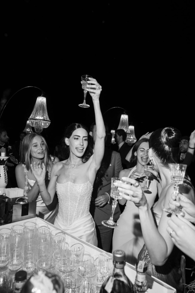 Eine Braut im Hochzeitskleid erhebt ein Glas und lächelt, umgeben von jubelnden Gästen bei einer lebhaften Hochzeitsfeier am Urlaubsort.