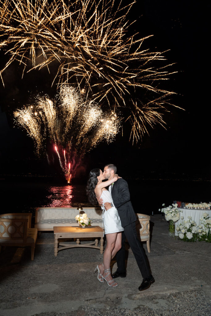 Ein Paar küsst sich nachts am Wasser, während am Himmel hinter ihnen ein Feuerwerk explodiert. Ein magischer Moment ihrer von Hochzeitsplaner Julia Weiß geplanten Destination Wedding.