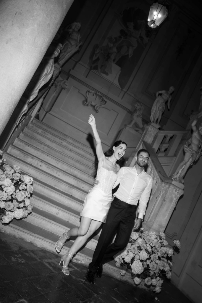 Ein glückliches Paar posiert auf einer verschnörkelten Treppe, umgeben von Blumen und klassischen Statuen, in Schwarz-Weiß - ein atemberaubender Moment, den Hochzeitsplaner Julia Weiß für eine Traumhochzeit am Urlaubsort eingefangen hat.