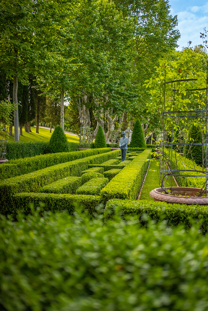Gepflegter Garten mit beschnittenen Hecken, einer Statue und hohen grünen Bäumen im Hintergrund unter blauem Himmel.
