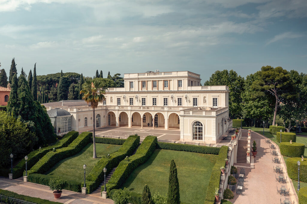 Große, elegante weiße Villa mit Bögen, gepflegten Hecken und Palmen unter einem teilweise bewölkten Himmel.