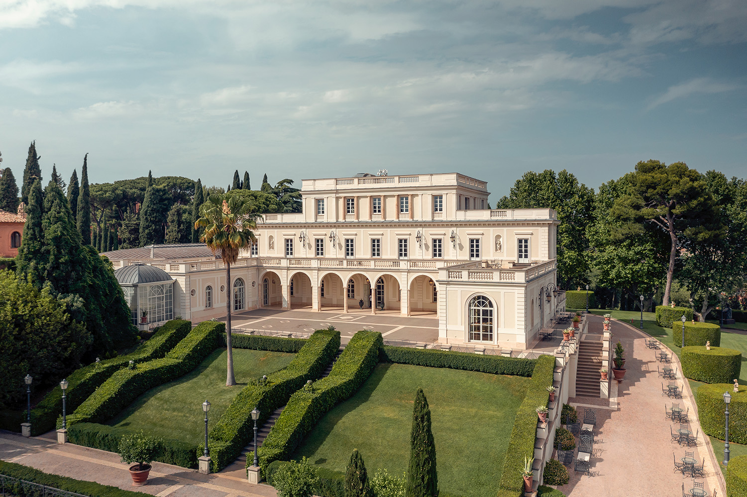 Große, elegante weiße Villa mit Bögen, gepflegten Hecken und Palmen unter einem teilweise bewölkten Himmel.