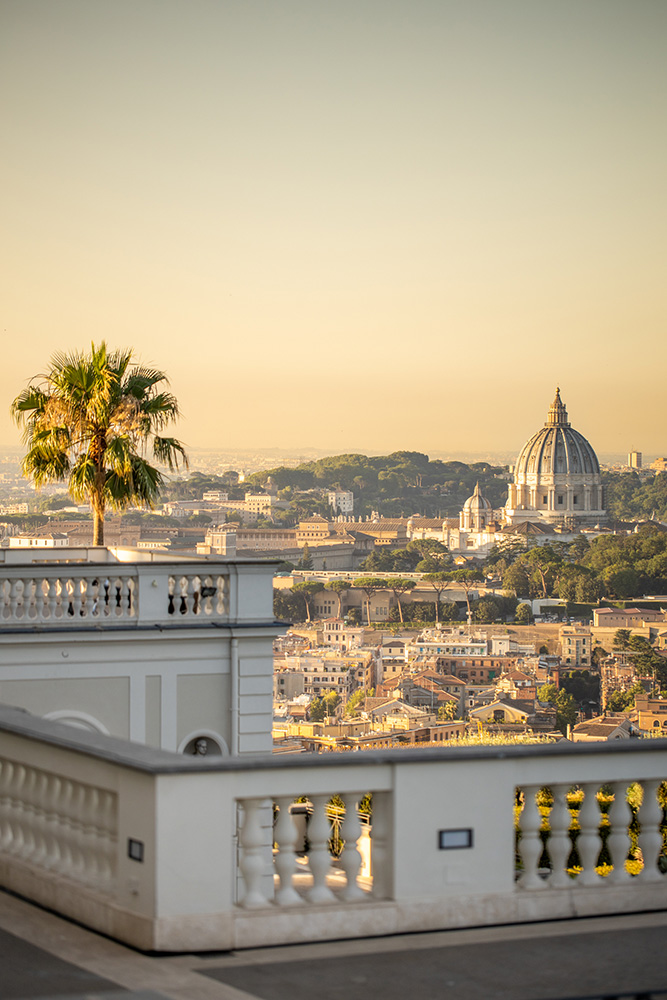 Panoramablick auf Rom mit einer Palme und der Kuppel des Petersdoms in der Ferne bei Sonnenuntergang.