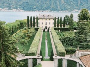 Elegante Villa mit gepflegten Gärten, hohen Zypressen und einem Springbrunnen inmitten einer Landschaft mit See und Bergen.