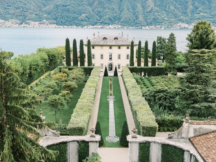 Elegante Villa mit gepflegten Gärten, hohen Zypressen und einem Springbrunnen inmitten einer Landschaft mit See und Bergen.